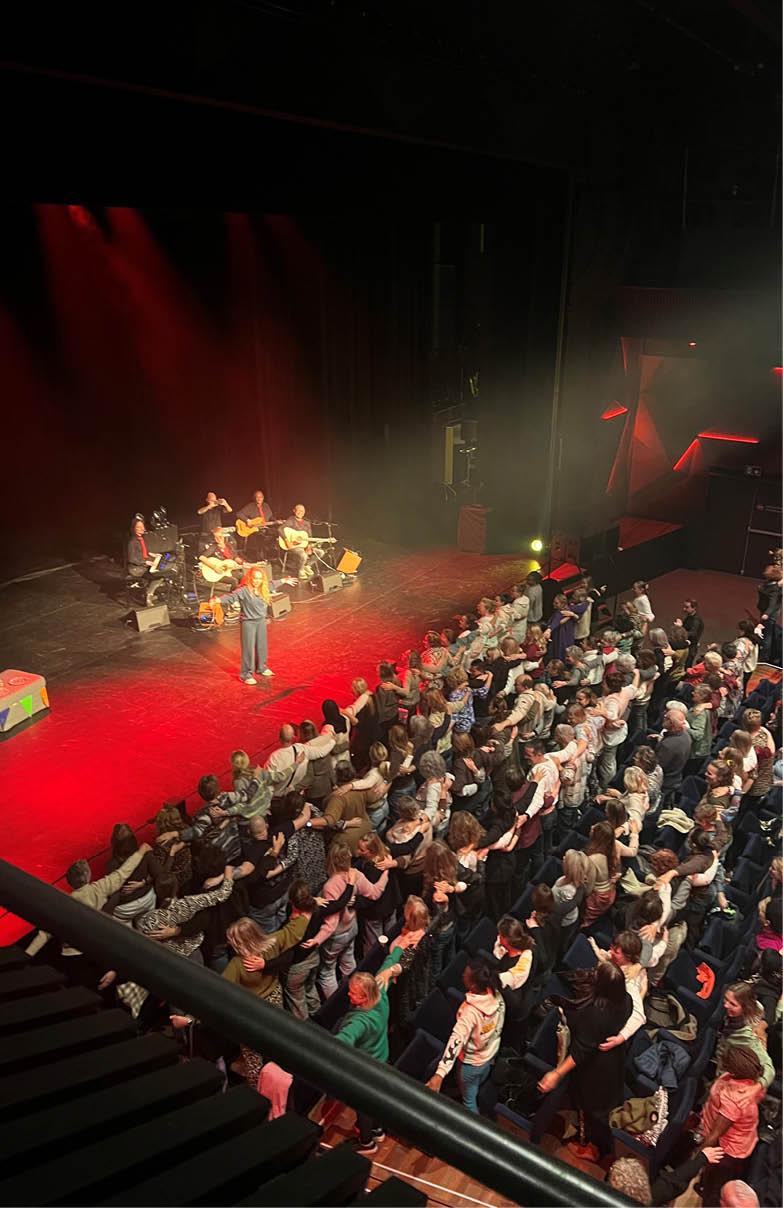A man is singing on stage in front of a large crowd of people. The audience is seated on chairs, and some are standing. The stage is illuminated by spotlights, and the man is wearing a tie. AI generated content