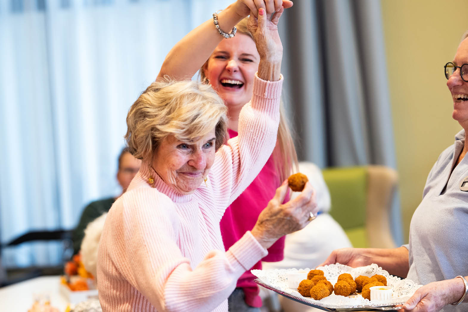 Three women are laughing and enjoying a meal together. They are holding food in their hands, with one woman holding a plate of food and the other two women holding food in their hands. The women are seated around a dining table, with a chair visible in the background. AI generated content